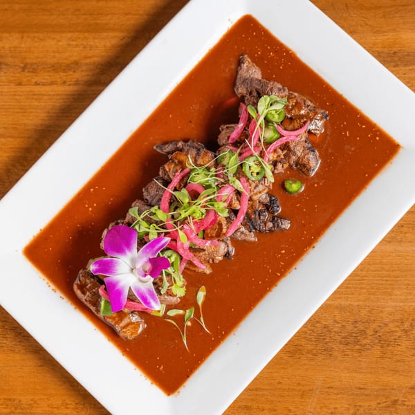 Grilled steak in a rich red sauce, garnished with pickled onions, jalapeños, and a purple orchid.